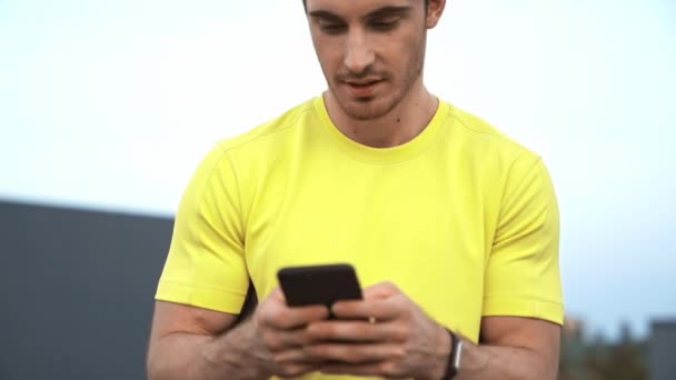 屋上に座っている黄色のスポーツウェアを着たスポーツマンの低角度の視点 手にスマートフォンを持ち テキストメッセージを送り 笑顔と真剣な顔をする — ストック動画