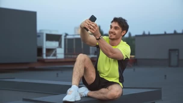 Przystojny Sportowca Siedzi Dachu Trzymając Smartfon Rękach Uśmiechając Się Grimace — Wideo stockowe