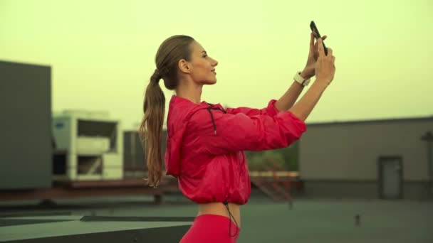 분홍색 스포츠웨어를 아름다운 스포츠우먼이 스마트폰을 포즈를 취하고 셀카를 옥상에 있습니다 — 비디오