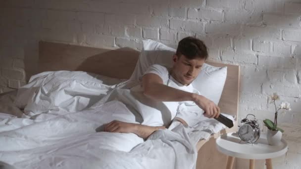 男人躺在毯子下在床上 拿着遥控器 改变电视节目 把遥控器放回去 — 图库视频影像