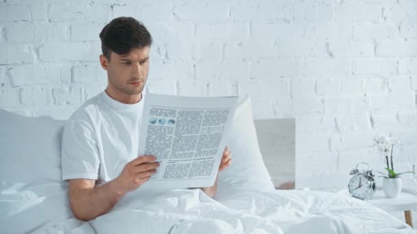 白いTシャツを着た男がベッドに横たわり 新聞を読み ページをめくる — ストック動画