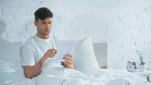 白いTシャツを着た男がベッドに横たわり デジタルタブレットを使い 朝に微笑む — ストック動画