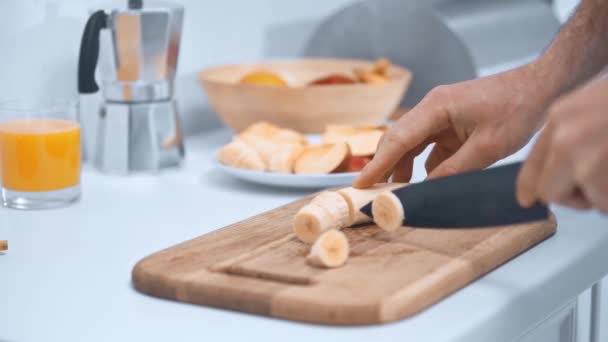 台所で木製のまな板にバナナを切る男のトリミングビュー — ストック動画