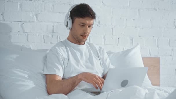 Szabadúszó Fehér Póló Hallgat Zenét Fejhallgató Dolgozó Laptop Míg Ágyban — Stock videók