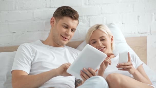 美しいカップルが話し 笑顔で ベッドの中でデジタルデバイスを使用しています — ストック動画