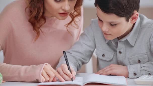 彼が机でコピーブックに書いている間 息子が宿題を手伝っている母親の見解をトリミング — ストック動画