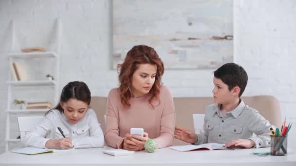 2人の十代の子供たちがテーブルで宿題をして 彼女がスマートフォンを使用している間 母親に助けを求める — ストック動画