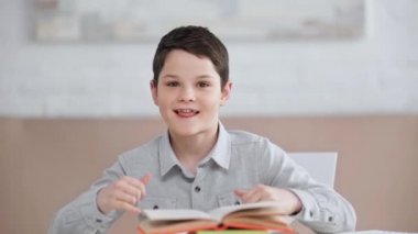 masada oturan gülümseyen preteen çocuk ön görünümü, başparmak yukarı ve kaya işaretleri gösteren, komik yüzler yapma ve baş kitap koyarak