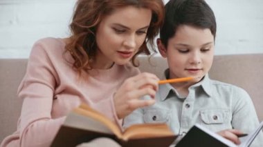 gülümseyen anne kitap tutan ve kanepede otururken ev ödevi ile oğlu yardım