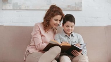 gülümseyen anne kanepede oturan ve kitap okurken oğlu ders kitabında yazma ve oturma odasında gülümseyen