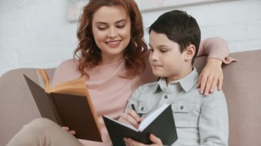 kanepede oturan gülümseyen anne, oğlu kucaklayan ve o ders kitabında yazarken kitap okuma
