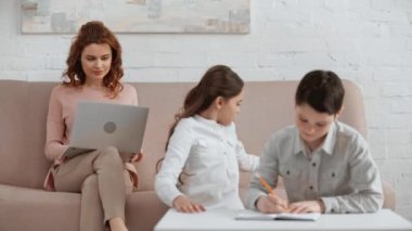 anne kanepede otururken ve dizüstü bilgisayar kullanırken birlikte ev ödevi yapan iki gülümseyen çocukların raf odak