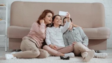 kanepe nin yanındaki halıda oturan, selfie çeken ve başparmak ve barış işaretleri gösteren mutlu gülümseyen aile