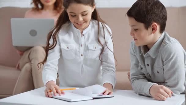 两个微笑的青春期前的孩子一起做作业 而母亲使用笔记本电脑在S台 — 图库视频影像