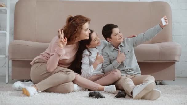 ソファの近くのカーペットに座って 自分撮りを取り 親指を上げて平和の兆候を見せる幸せな笑顔の家族 — ストック動画