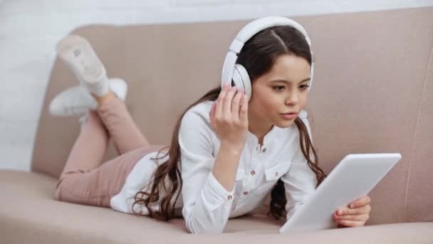 开朗的青春期前孩子躺在桌子上 在耳机里听音乐 使用数字平板电脑唱歌 — 图库视频影像