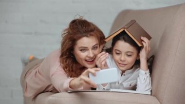 neşeli preteen çocuk kafasına kitap koyarak, anne ile selfie alarak ve evde kanepede yatarken gülümseyerek