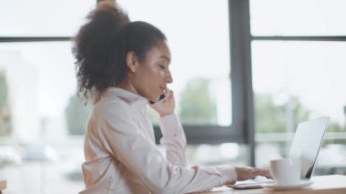 afro-amerikan iş kadını akıllı telefon konuşuyor ve ofiste dizüstü bilgisayar kullanarak