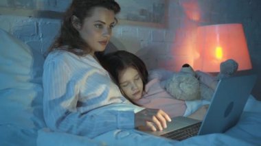 oyuncak ayı ile uyuyan kızı yakınında dizüstü bilgisayar kullanan anne