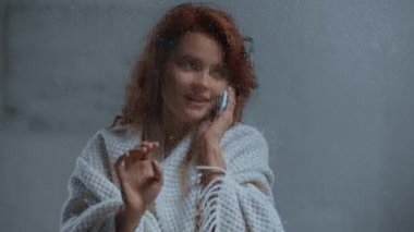 Akıllı telefondan konuşan gülümseyen kadın camlı pencerenin yanında yağmur damlalarıyla konuşuyor.