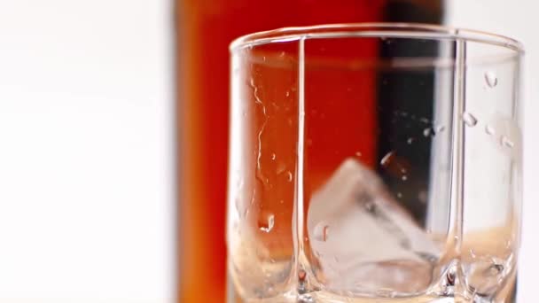 白で隔離されたウイスキーの瓶の近くのガラスに氷のキューブが落ち — ストック動画