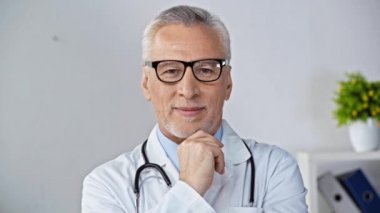 Gözlüklü olgun bir doktor gülümsüyor, çenesine dokunuyor ve el sallıyor.