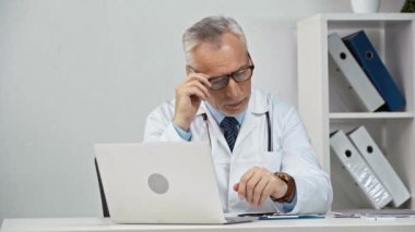 Olgun doktor reçete yazıyor, gözlüğünü çıkarıyor ve şakaklarına dokunuyor. 