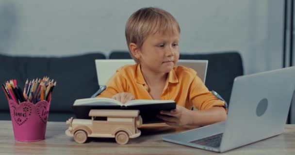 Ładny Chłopiec Czytanie Książki Pobliżu Laptopa Podczas Learning Domu — Wideo stockowe