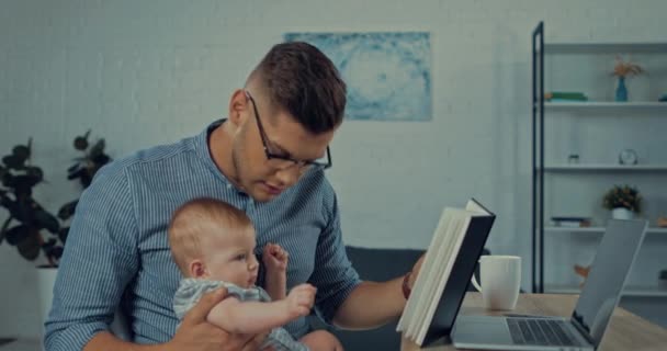 父亲戴着眼镜 在笔记本电脑旁抱着婴儿儿子和笔记本 — 图库视频影像
