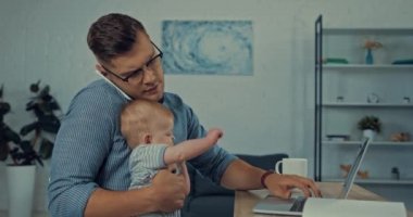 Yakışıklı baba akıllı telefonla konuşurken dizüstü bilgisayar kullanıyor ve kucağında bebek var.