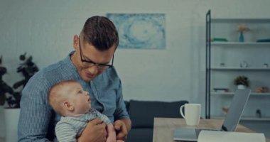 Baba kucağında bebek varken dizüstü bilgisayarını işaret ediyor.