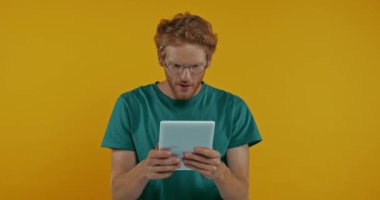 Sarı üzerine izole edilmiş dijital tablet kullanan gözlüklü, şok olmuş kızıl saçlı adam.