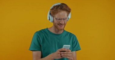 Kulaklıklı mutlu kızıl adam sarıda izole edilmiş akıllı telefonuyla dans ediyor.