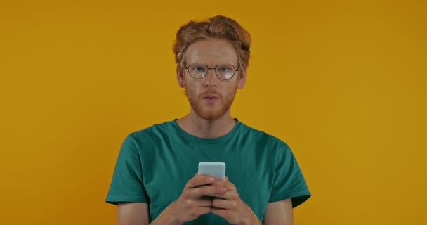Verrast Roodharige Man Bril Met Behulp Van Smartphone Geïsoleerd Geel — Stockvideo