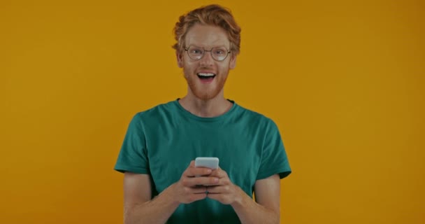 Glad Rödhårig Man Glasögon Pekar Smartphone Isolerad Gul — Stockvideo