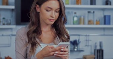 Önlüklü genç bir kadın mutfakta akıllı telefon kullanıyor. 