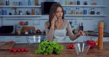 Önlüklü kadın masadaki malzemelerin yanında akıllı telefondan konuşuyor.