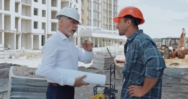 建設現場で話すハード帽子の青写真とビルダーを持つエンジニア — ストック動画