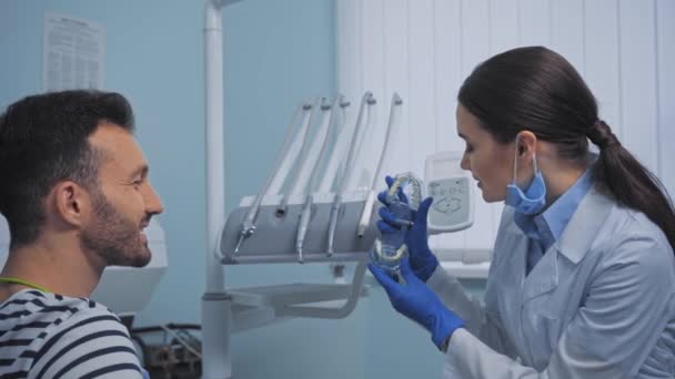 Pacjent Patrzący Uśmiechniętego Dentystę Pokazującego Model Zębów Klinice — Wideo stockowe