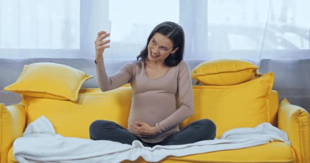 妊娠中の女性持っていますビデオ通話のスマートフォン上のソファ — ストック動画