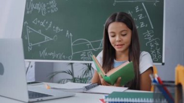 Çocuk dizüstü bilgisayarın yanında yüksek sesle kitap okuyor ve masanın üstünde defterler var.