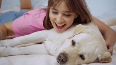 Genç bir kadın yatak odasında battaniyeye uzanıp Golden Retriever köpeğine sarılıyor.