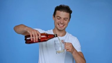 Beyaz tişörtlü mutlu adam maviden izole edilmiş bardağa bira döküyor.