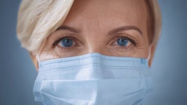 Medikal maskeli mavi gözlü orta yaşlı kadın kameraya bakıyor.