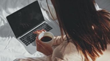 Kahve içen ve dizüstü bilgisayarla yatakta yazan serbest çalışan birinin genel görünümü.