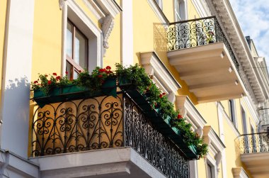 Çiçeklerle süslenmiş siyah süslü ferforje balkon. Kırmızı sardunyalı kutular. Alt taraf görünümü