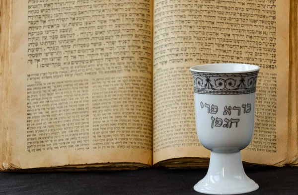 Otwarta stara Biblia żydowska i biała porcelanowa filiżanka wina kiddush na czarnym tle. Otwarta strona z pism świętych. Napis na filiżance w języku hebrajskim: twórca owocu winorośli. Selektywna koncentracja — Zdjęcie stockowe