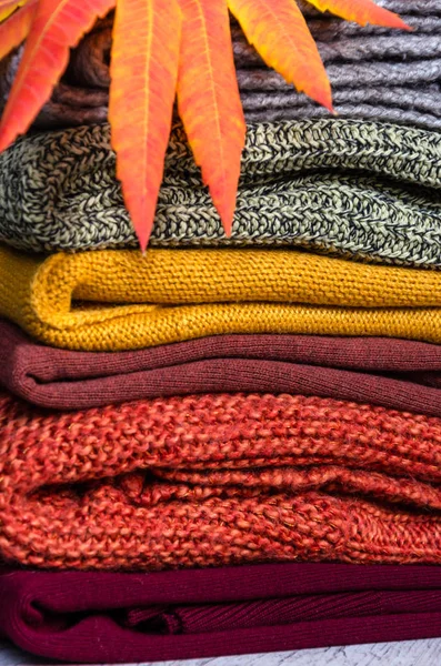 Stack of folded warm knitted women's sweaters in warm colors and bright autumn leaves. Closeup — Stock Photo, Image