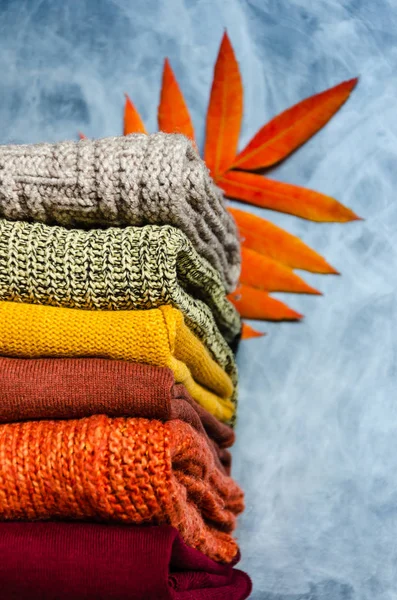 Montón de suéteres de punto tibios doblados de las mujeres en colores cálidos y hojas de otoño brillantes sobre fondo gris. Primer plano. Copiar espacio — Foto de Stock