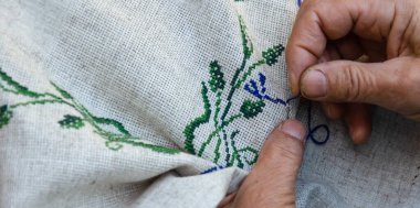 Yaşlı bir kadının elleri keten kumaşa çapraz dikişli çiçek deseni işliyor. Nakış, el işi, iğne konsepti. Yakın çekim.